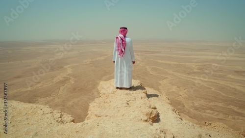 Adult man in traditional Arabian clothing stands on the edge of a cliff in the desert. Saudi Arabia