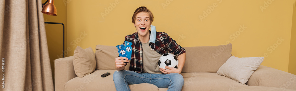 horizontal image of excited man in medical mask holding football and sport match tickets near remote controller, end of quarantine concept horizontal image of excited man in medical mask holding football and sport match tickets near remote controller, end of quarantine concept