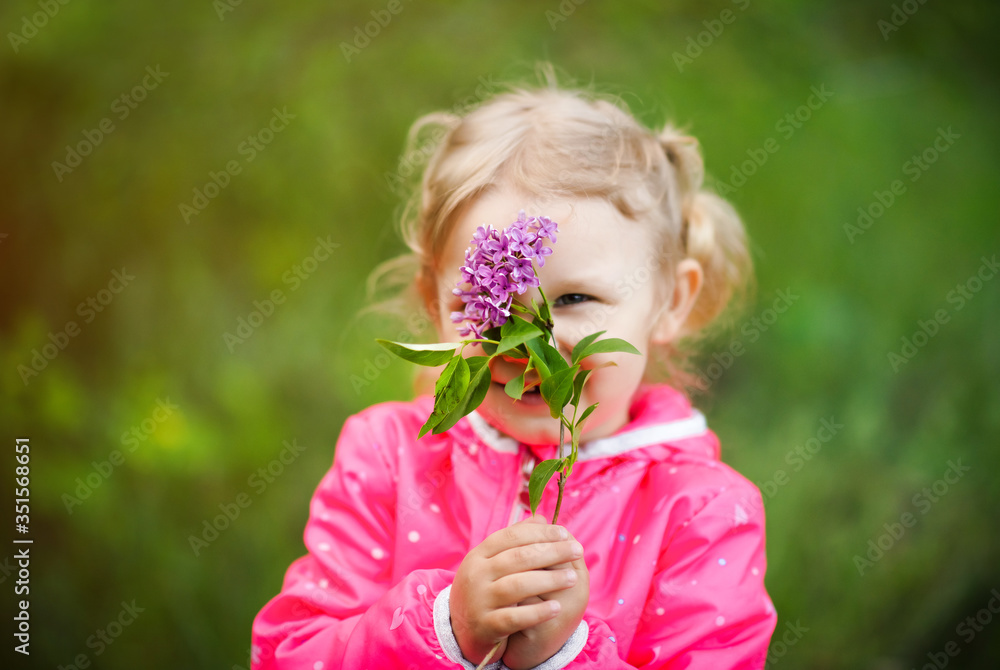 Portrait of little 3 years old blonde girl in pink jacket hiding behind branch of purple lilac in hand. Lilac and unrecognizable smiling kids face. Blossom spring concept Portrait of little 3 years old blonde girl in pink jacket hiding behind branch of purple lilac in hand. Lilac and unrecognizable smiling kids face. Blossom spring concept