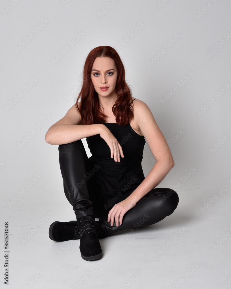 Portrait of a pretty girl with red hair wearing black leather pants, shirt and boots. full length sitting pose, isolated against a studio background Portrait of a pretty girl with red hair wearing black leather pants, shirt and boots. full length sitting pose, isolated against a studio background