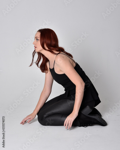 Portrait of a pretty girl with red hair wearing black leather pants, shirt and boots. full length sitting pose, isolated against a studio background
