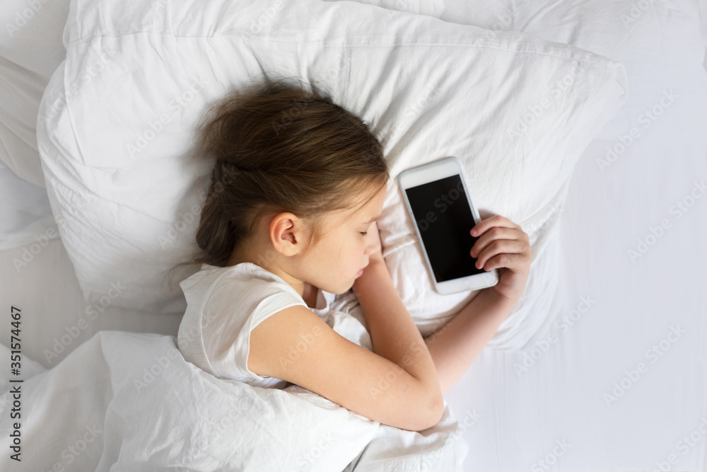 Tired Caucasian girl child sleeps with a phone on a white pillow, top view, the concept of children and gadgets, child health and child safety online Tired Caucasian girl child sleeps with a phone on a white pillow, top view, the concept of children and gadgets, child health and child safety online