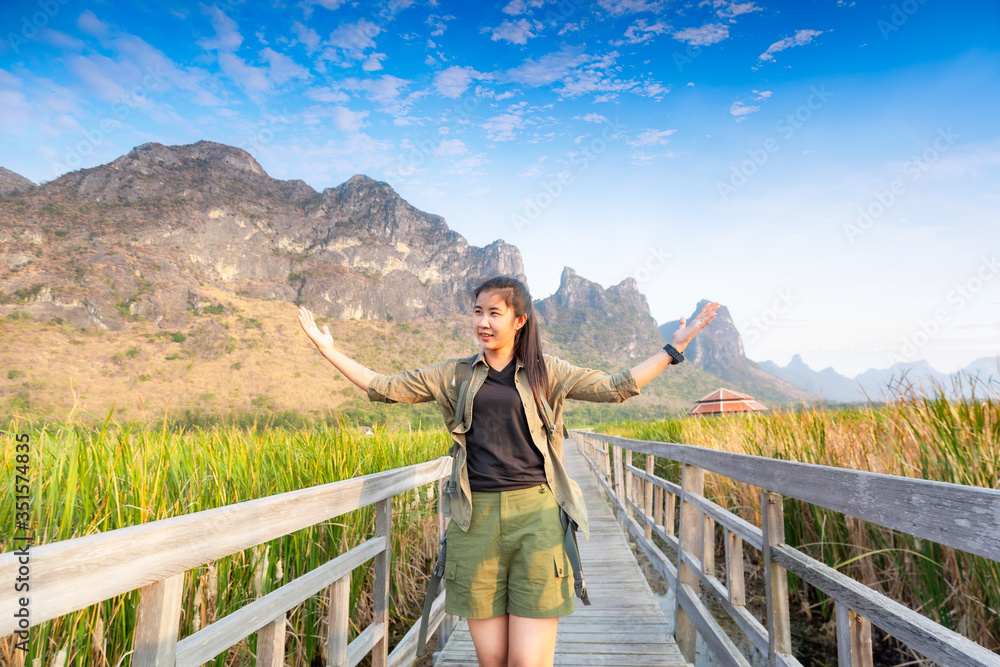 Naklejka premium Asian hikers carry heavy backpacking on a small Pavilion outdoor hiking path on a wooden bridge in a swamp with meadows with a blue mountain background. Khao Sam Roi Yot National Park