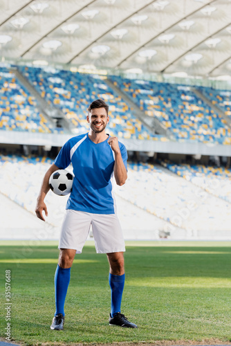 happy professional soccer player in blue and white uniform with ball showing yes gesture on football pitch at stadium © LIGHTFIELD STUDIOS
