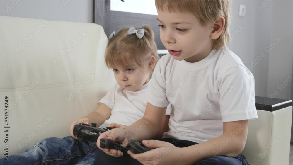 Blonde-haired children a girl and a boy play a game console. Happy children play sitting on a white sofa at home. Blonde-haired children a girl and a boy play a game console. Happy children play sitting on a white sofa at home.