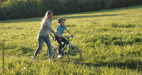 Mother teaching son to ride bicycle. Happy cute boy in helmet learn to riding a bike in park on green meadow in summer day at sunset time. Family weekend. 4K video Slow motion