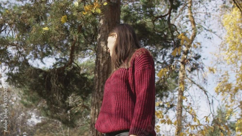 Young attractive woman with brown hair in red knitted jumper standing in sunlit autumn forest. Low angle 4K 360 degree tracking arc shot.