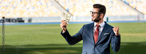 smiling young businessman in suit and glasses with money showing yeas gesture at stadium, panoramic shot photo