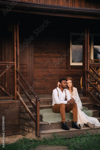 Boho style wedding. Caucasian bride and african groom sit embracing on the porch of the house.