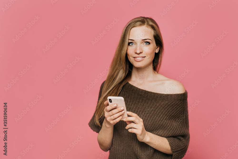 Beautiful young redhead woman dressed in brown knitted sweater with open shoulder, holds her smartphone, cute smiling, satisfied by talk with her boyfriend, posing over pink background with copy space Beautiful young redhead woman dressed in brown knitted sweater with open shoulder, holds her smartphone, cute smiling, satisfied by talk with her boyfriend, posing over pink background with copy space