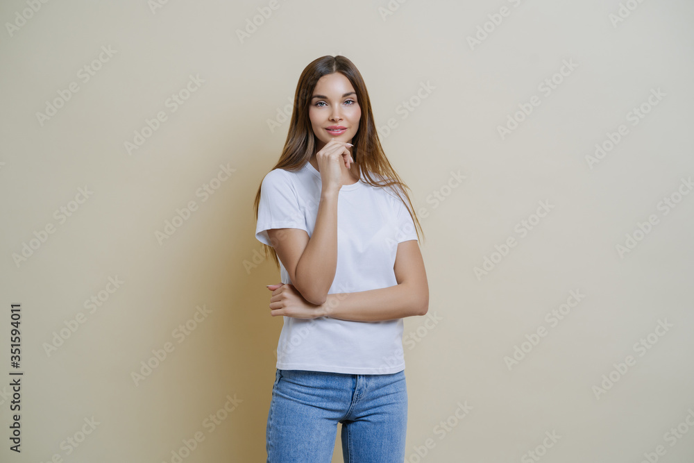 Photo of brunette young European woman holds chin, wears white t shirt and jeans, enjoys conversation with her mates, isolated over brown background. People, lifestyle, face expressions concept Photo of brunette young European woman holds chin, wears white t shirt and jeans, enjoys conversation with her mates, isolated over brown background. People, lifestyle, face expressions concept