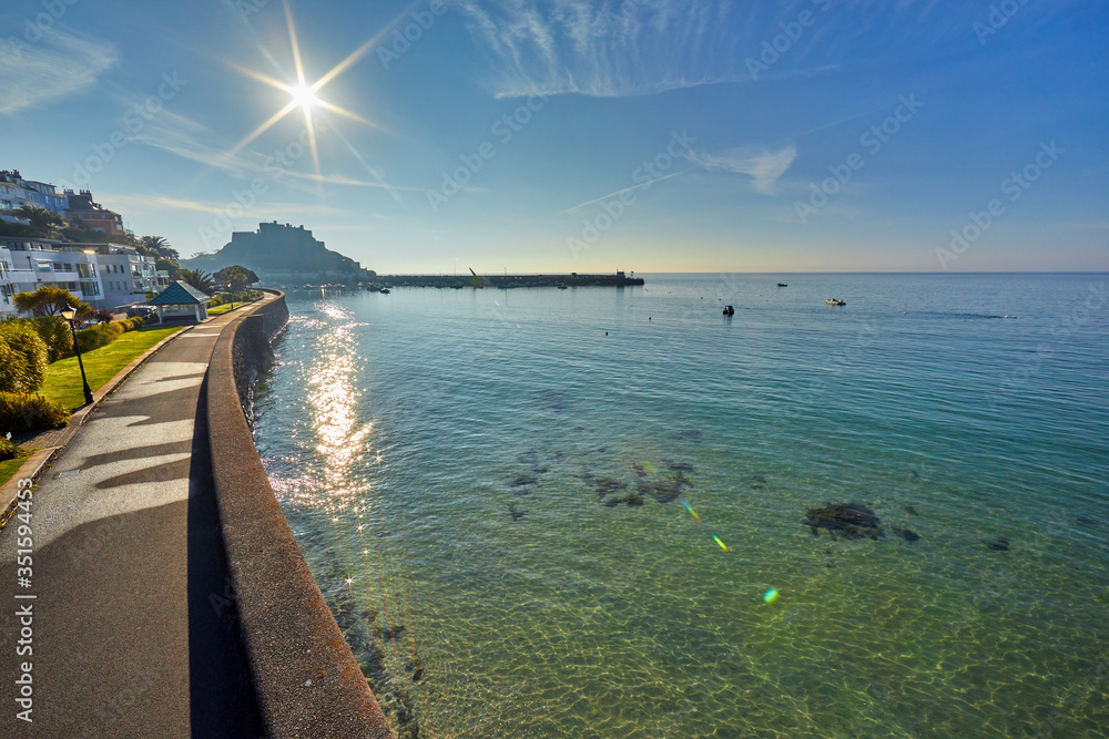 Obraz premium Image of Gorey promenade with Gorey Castle and harbour in the background at high tide with a blue sky and early morning sunshine. Jersey, Channel Islands, Uk