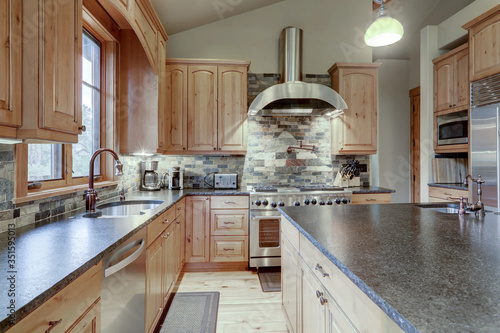 Large luxury wooden kitchen interior with light wood hardwood floor and dark granite slab on island with huge windows.