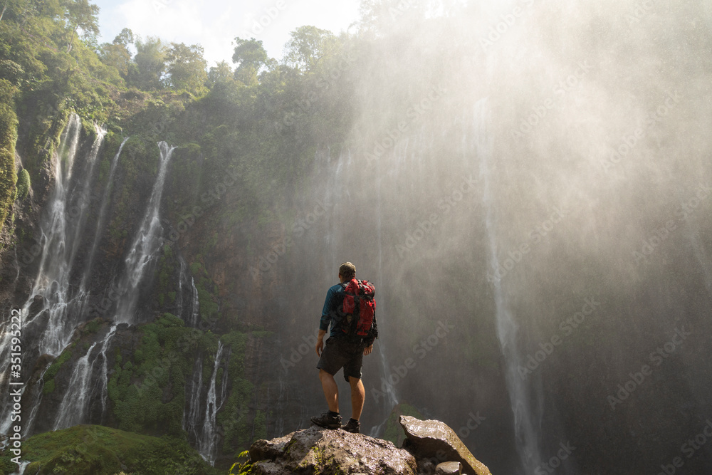 A backpacking adventurer gazes in awe at the massive rock circus formed by the intense waterfalls at Tumpak Sewu in East Java, Indonesia.