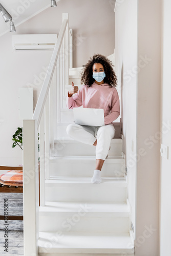 curly african american freelancer in medical mask sitting on stairs and showing thumb up near laptop