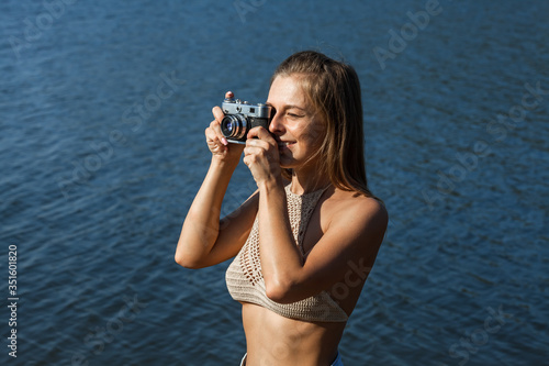 Young white girl with blonde hair holding a camera and taking pictures on the background of the sea, and she smiles. Summer sunny day.