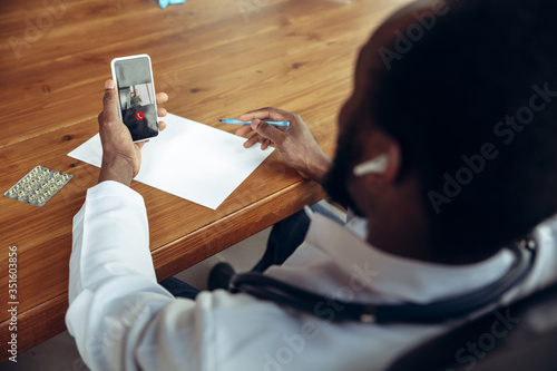 Doctor advising the patient online with smartphone. African-american doctor during his work with patients, explaining recipes for drug. Daily hard work for health and lives saving during epidemic. photo