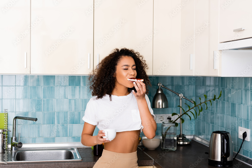 cheerful african american girl eating toast bread with jam and holding cup of coffee cheerful african american girl eating toast bread with jam and holding cup of coffee