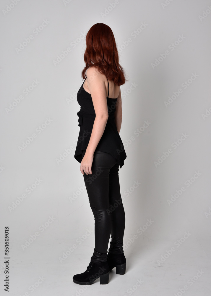 portrait of a pretty girl with red hair wearing black leather pants and top. Full length standing pose isolated against a grey studio background portrait of a pretty girl with red hair wearing black leather pants and top. Full length standing pose isolated against a grey studio background