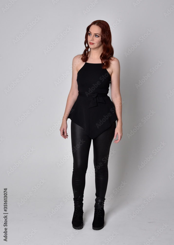 portrait of a pretty girl with red hair wearing black leather pants and top. Full length standing pose isolated against a grey studio background portrait of a pretty girl with red hair wearing black leather pants and top. Full length standing pose isolated against a grey studio background