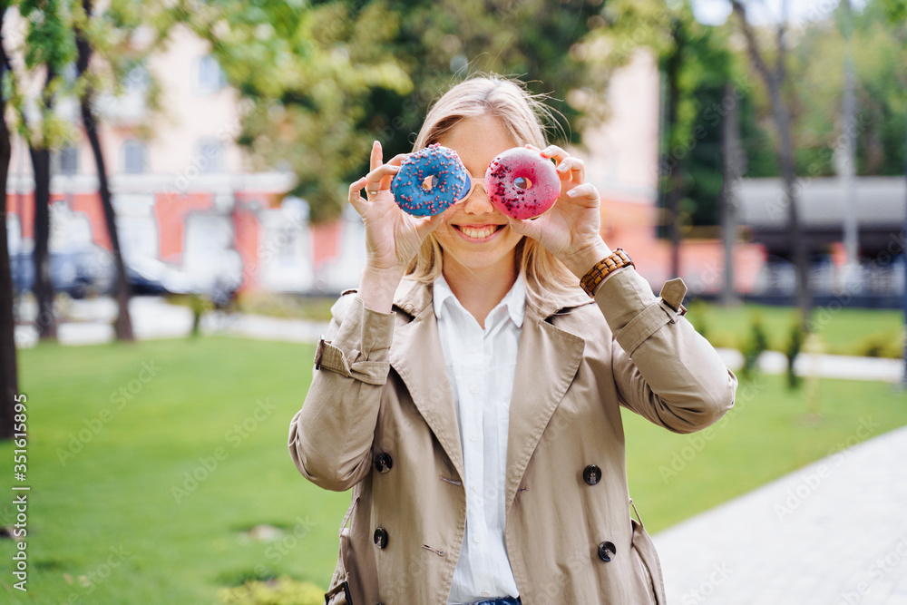 Beautiful, attractive blonde girl in a beige cloak and blue jeans is fooling around with donuts in her hands. Woman holding donuts near the face instead of eyes and smiling broadly. 
Looking through