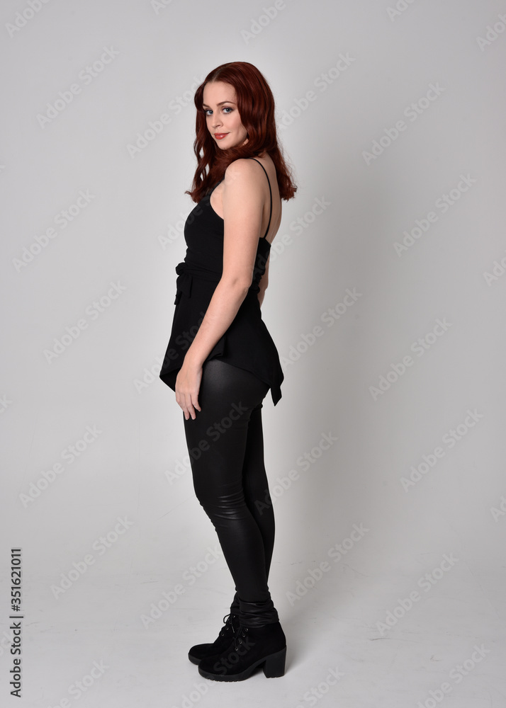 portrait of a pretty girl with red hair wearing black leather pants and top. Full length standing pose in side profile isolated against a grey studio background portrait of a pretty girl with red hair wearing black leather pants and top. Full length standing pose in side profile isolated against a grey studio background