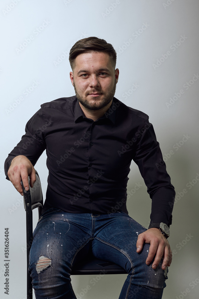 Young Bearded Dark Haired Man In Black Stylish Shirt, On Gray Background, Confident Serious Man Sitting On Chair And Looking Straight. Concept Of Ordinary Ordinary People Who Work In An Office Young Bearded Dark Haired Man In Black Stylish Shirt, On Gray Background, Confident Serious Man Sitting On Chair And Looking Straight. Concept Of Ordinary Ordinary People Who Work In An Office