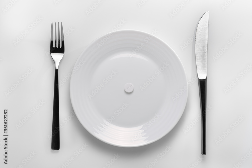 Table setting with a white plate filled with  white pills next to cutleries on a white background. To represent a concept of pharmaceutical addiction, healthy nutrition, pharmaceutical industry
