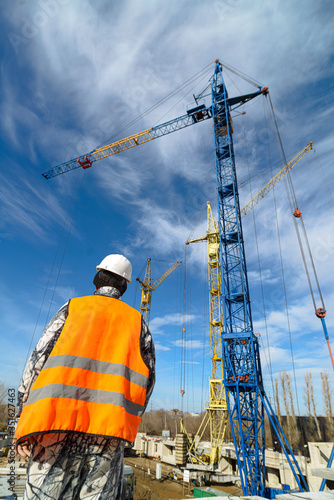 A worker in overalls looks at the high-rise construction cranes on the construction site against a blue sky.
