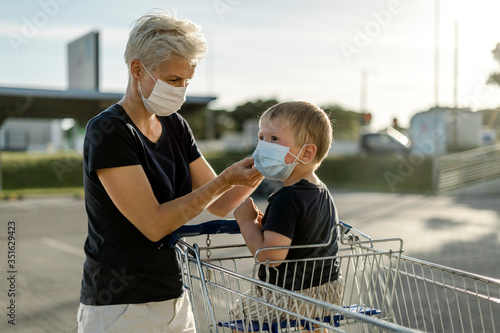 Mother helping her son sitting in a shopping car to wear a protective mask.