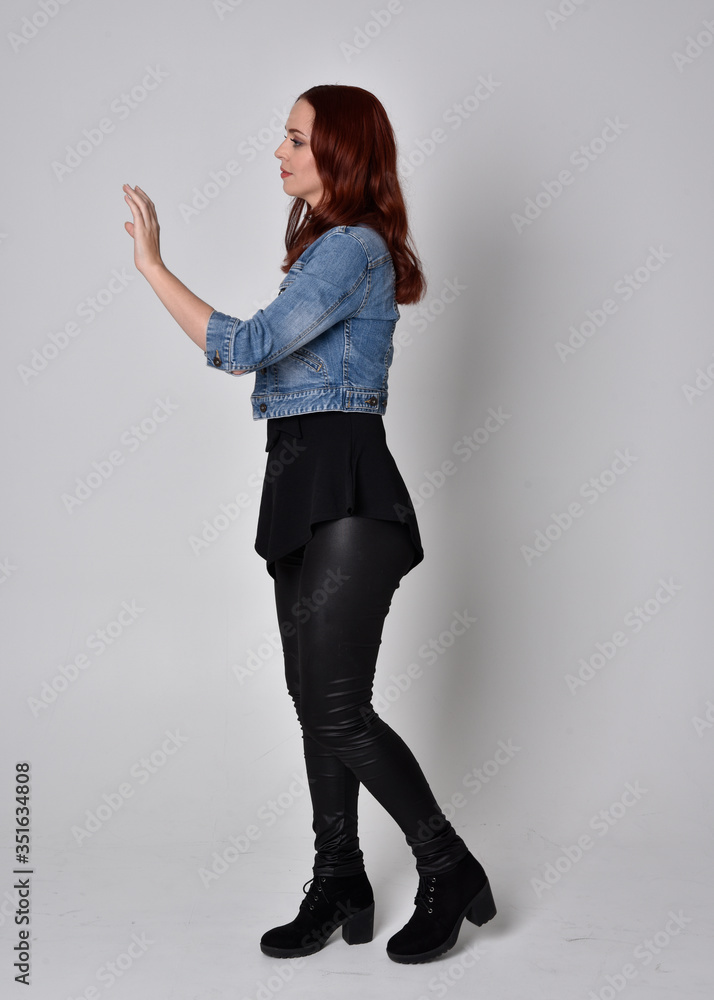 portrait of a pretty girl with red hair wearing black leather pants and denim jacket. Full length standing pose in side profile isolated against a grey studio background portrait of a pretty girl with red hair wearing black leather pants and denim jacket. Full length standing pose in side profile isolated against a grey studio background