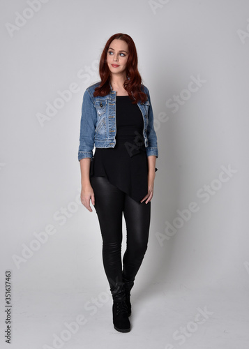  portrait of a pretty girl with red hair wearing black leather pants and top. Full length standing pose isolated against a grey studio background