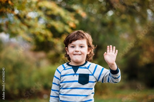 Summer, childhood, leisure, human gesture concept - happy little boy waving hand. Portrait of a little boy in the woods waving his hand. Portrait of a boy.