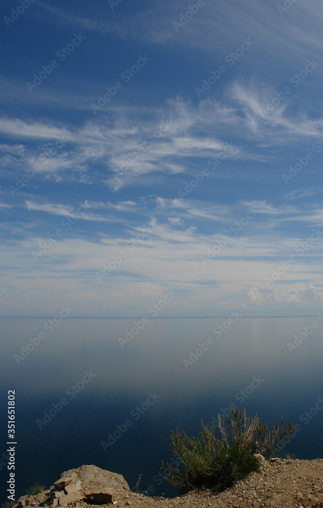 The clouds over the calm water of Baikal Lake on a clear summer day, view from the rocky shore of Cape Khoboy (Siberia, Russia). Wild mugwort (Artemisia monostachya), growing on the edge of a cliff