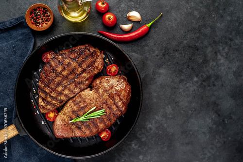 Two grilled beef steaks in a pan with spices on a stone background