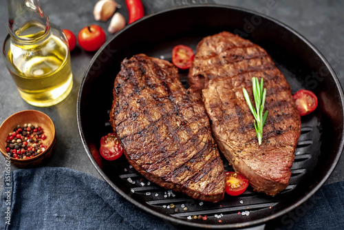 Two grilled beef steaks in a pan with spices on a stone background