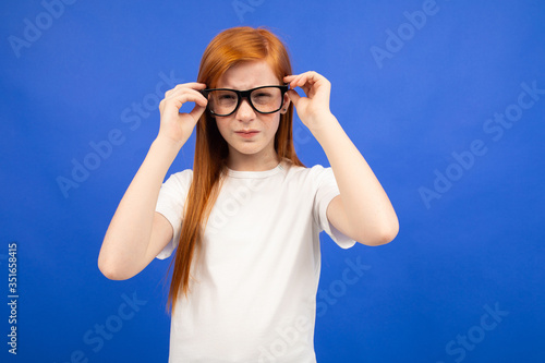 red-haired teenager girl squints while holding glasses in her hand on a blue studio background