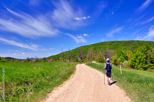 Tourism and outdoor activities. A woman with a backpack on her back is walking along a dusty country road, a blue sky in the background.