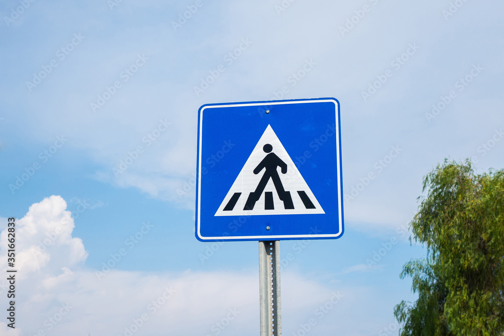 International traffic sign 'Pedestrian crossing' on clouded blue sky background. Symbolic person crosses street from right to left