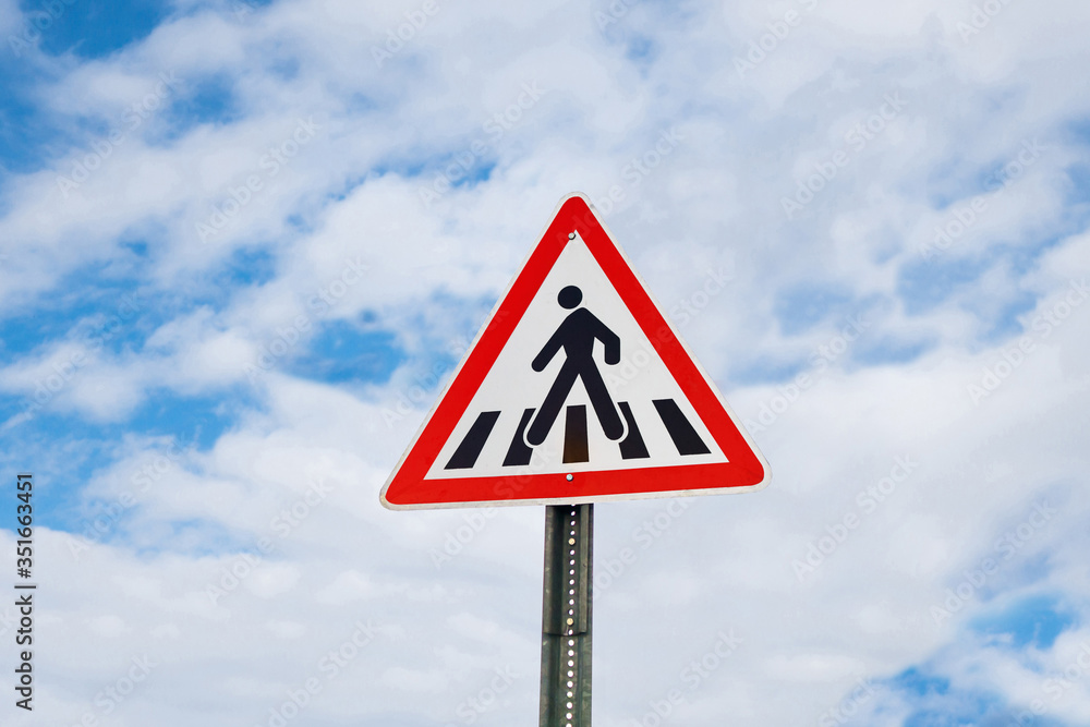 International warning traffic sign 'Pedestrian crossing' on metal pillar. Cloudy blue sky is on background