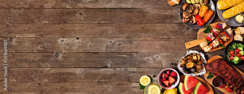 Summer BBQ or picnic food corner border over a rustic wood banner background. Assorted grilled meats, vegetables, fruits, salad and potatoes. Overhead view with copy space. photo