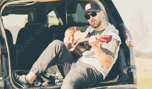 Man from the milenials generation sits in the trunk of a car and plays an acoustic guitar - relax and get out of town - playing the guitar while traveling - strong sunlight photo