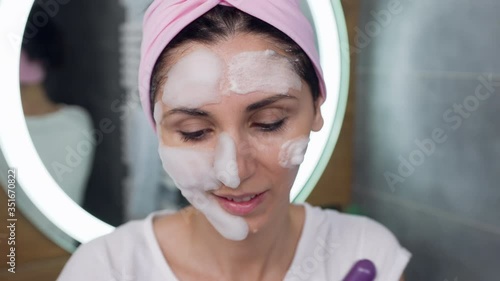 Portrait of charming joyful young brunette in headband which cleaning her face with cosmetic foam and posing on camera photo