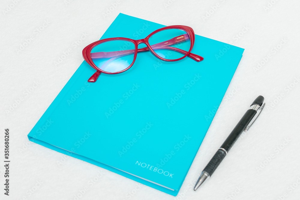 Blue notebook, pen and red women's glasses, white background, top view, copy space Blue notebook, pen and red women's glasses, white background, top view, copy space