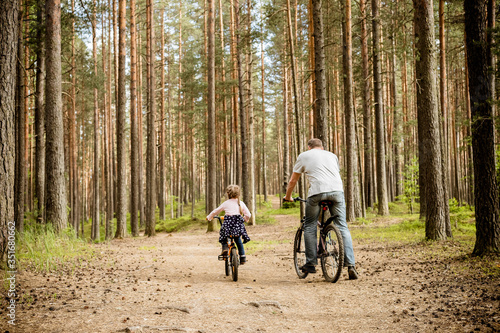 back view of father and daughter riding bicycles around forest.Young family in summer cycling in park.The theme family sports outdoor recreation.Adventure leisure concept.