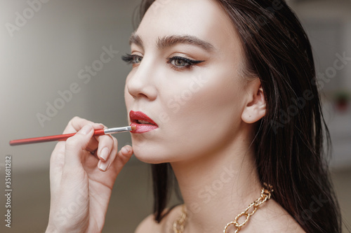 professional makeup artist with brush applies lip gloss to her client woman in beauty salon