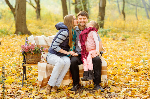 Dad, mom and their daughter are sitting on a bench in the autumn park. Parents with a little girl took refuge in a blanket to keep warm.