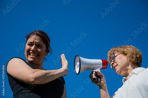 The conflict of generations. An emotional elderly woman shouting at her daughter in a megaphone. An elderly mother swears at a middle-aged woman on a loudspeaker on a blue background. © Михаил Решетников