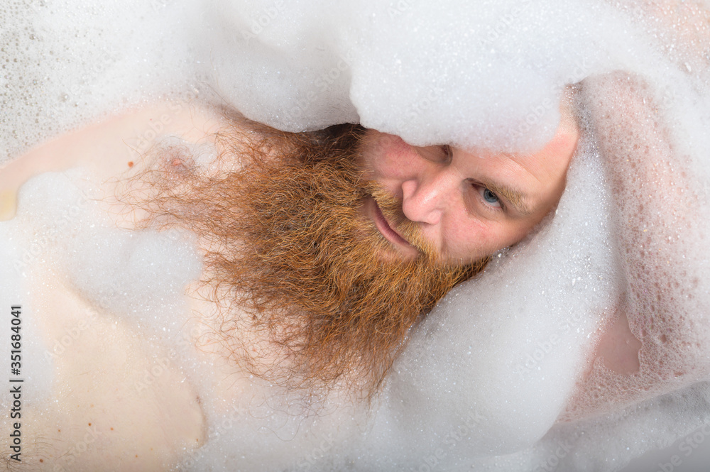 Portrait of a bald man with a long red beard takes a bath with foam. Top view on a cheerful funny guy in soapy water. Portrait of a bald man with a long red beard takes a bath with foam. Top view on a cheerful funny guy in soapy water.