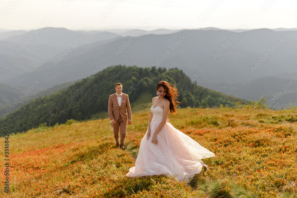 The bride and groom are standing close to each other against the backdrop of autumn mountains. The groom admires his wife.